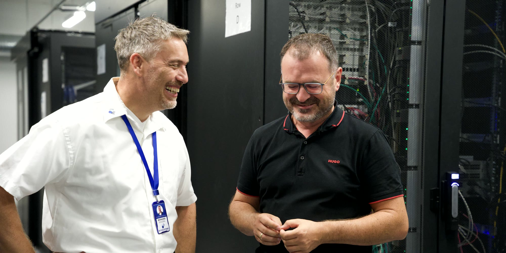Wolfgang Asbeck und Bernhard Schlögl lachen gemeinsam zwischen den Serverschränken im Rechenzentrum München.